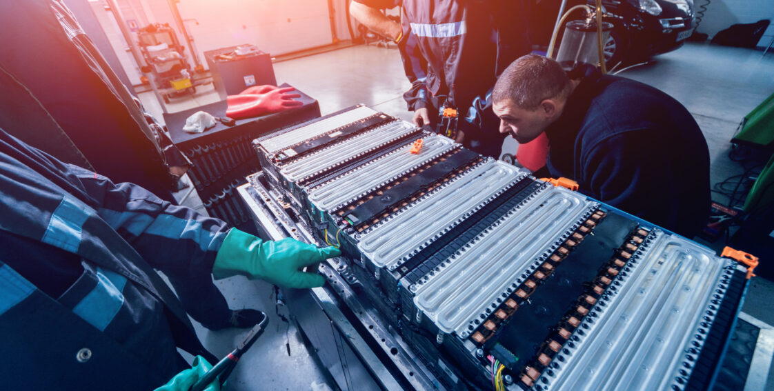 Three technicians are standing next to an electric vehicle battery that is on a table. One worker is crouched down in front of it to examine it. Another worker stands behind the crouched technician with his hand on his hip, surveying the scene. The third employee is standing on the other side of the table wearing insulated gloves and holding insulated pliers.