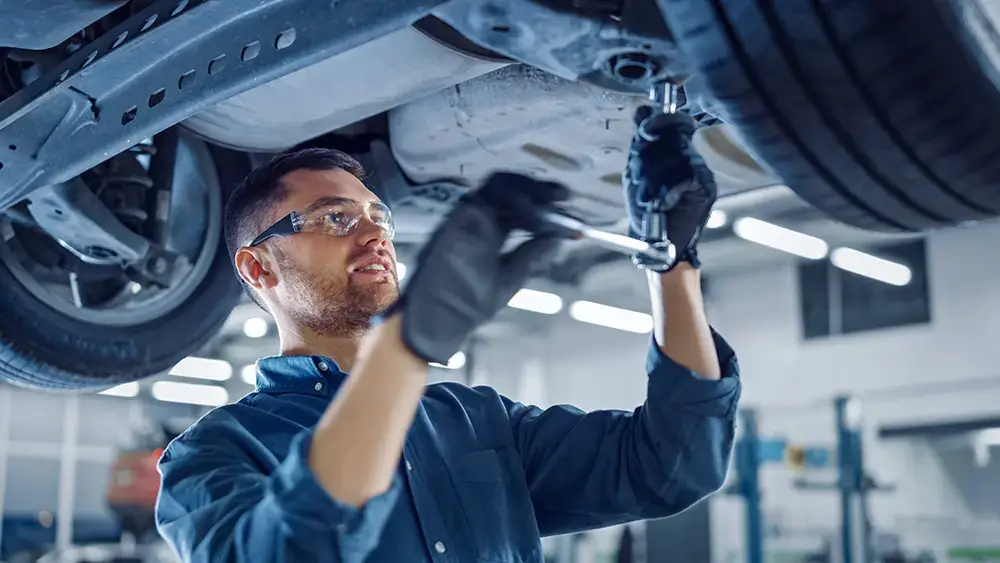 Portrait Shot of a Handsome Mechanic Working on a Vehicle in a Car Service. Professional Repairman is Wearing Gloves and Using a Ratchet Underneath the Car. Modern Clean Workshop.
